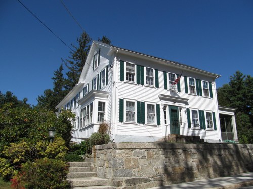 A white colonial-style house with green shutters, steps leading up, and an American flag, surrounded by trees and shrubs.
