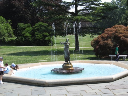 A decorative fountain with a statue in the center, surrounded by greenery and people enjoying the park.