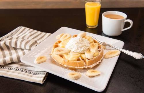 A plate of waffles topped with banana slices and whipped cream, accompanied by orange juice and a cup of coffee.