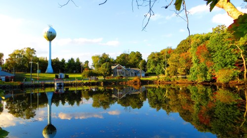 A serene lake reflects a water tower and colorful trees under a clear blue sky.
