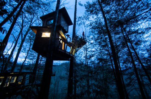A treehouse perched high in trees, illuminated at dusk, surrounded by dense forest and mountains in the background.