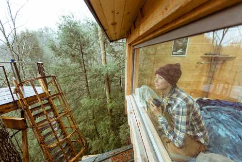 A person sits by a window in a treehouse, gazing outside at a misty forest while holding a cup.