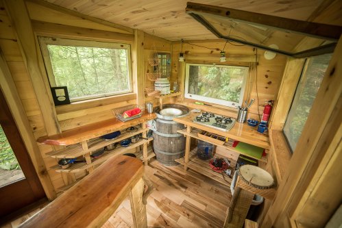 Cozy wooden kitchen with a barrel sink, gas stove, and large windows overlooking a forested area.