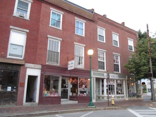 A brick building with shops on the ground floor and residential windows above, set in a quaint street scene.