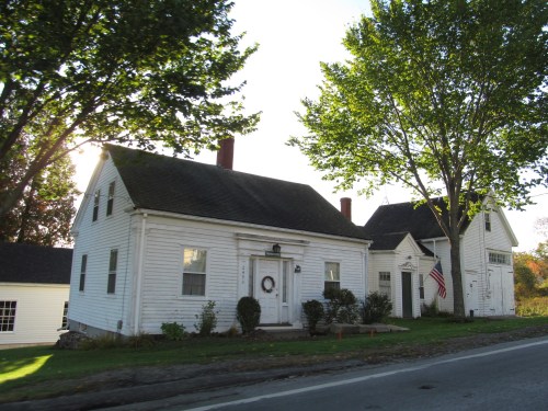 Two white houses with green lawns and large trees, one displaying an American flag, along a quiet road.