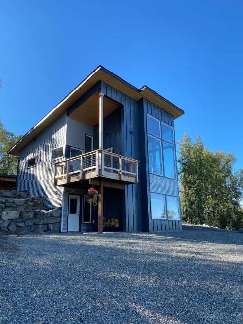 Modern two-story house with large windows, a balcony, and a gravel driveway, surrounded by trees.