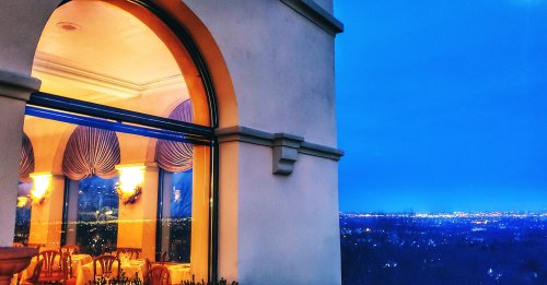A view of a beautifully lit dining area through an arched window, overlooking a city skyline at dusk.