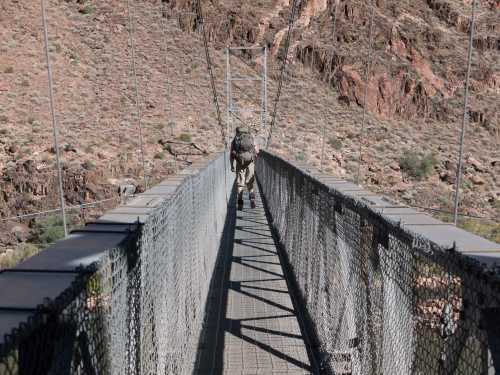 A hiker crosses a suspension bridge over a canyon, surrounded by rocky terrain and sparse vegetation.