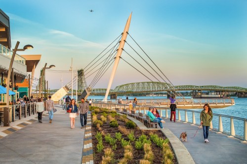 A scenic waterfront walkway with people, a bridge in the background, and a modern sculpture under a clear sky.