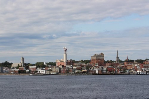 A coastal city skyline with various buildings, a tower, and church spires under a cloudy sky, viewed from the water.