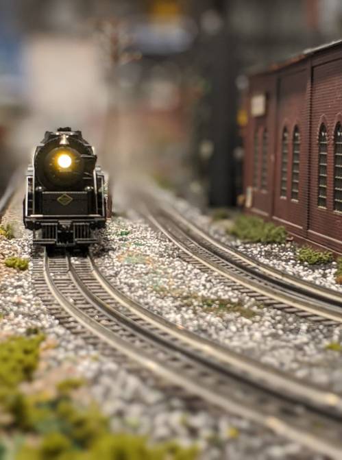 A model steam train approaches on a detailed miniature railway, surrounded by tracks and a brick building.