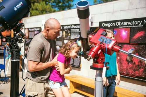 A man helps a girl look through a telescope at an observatory, with educational displays in the background.