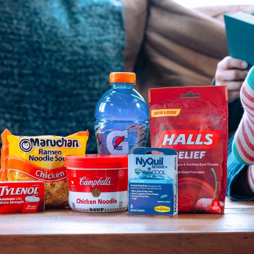 A cozy scene with a blue sweater, a bowl of ramen, Gatorade, soup, cold medicine, and cough drops on a wooden table.