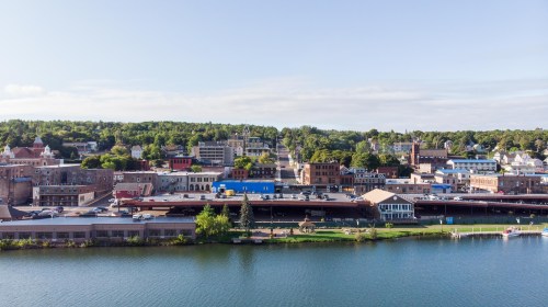 A scenic view of a waterfront town with buildings, trees, and a calm river under a clear blue sky.
