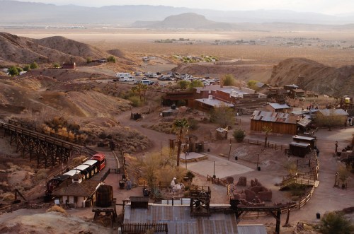 Aerial view of a desert town with rustic buildings, mountains in the background, and a train track winding through.