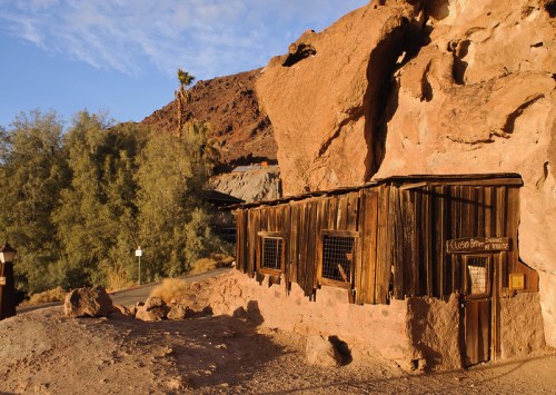 A rustic wooden structure built into a rocky landscape, surrounded by desert vegetation and mountains in the background.