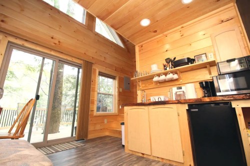 Cozy wooden cabin interior with a kitchen, large windows, and a sliding door leading to a deck. Natural light fills the space.