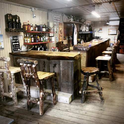 Rustic bar interior with wooden stools, shelves of liquor, and a cozy, inviting atmosphere.