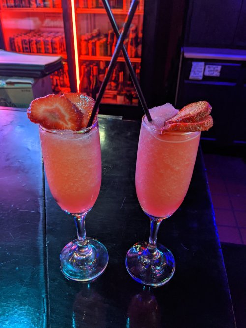 Two pink cocktails in tall glasses, garnished with strawberries and served with black straws, on a dark bar surface.