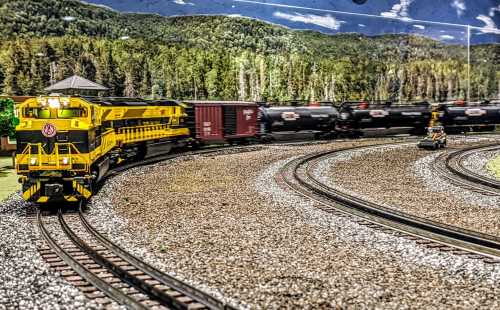 A model train set featuring a yellow locomotive and freight cars on curved tracks, surrounded by a scenic backdrop of trees.