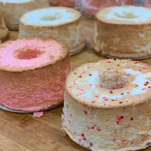 A variety of round, layered cakes with pink and white frosting, some with a hole in the center, on a wooden surface.