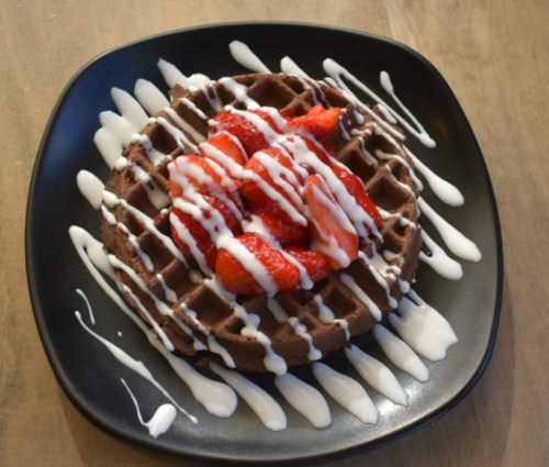 A chocolate waffle topped with fresh strawberries and drizzled with white icing on a black plate.