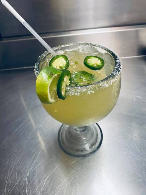 A refreshing cocktail in a large glass, garnished with lime and jalapeño slices, with a salted rim.
