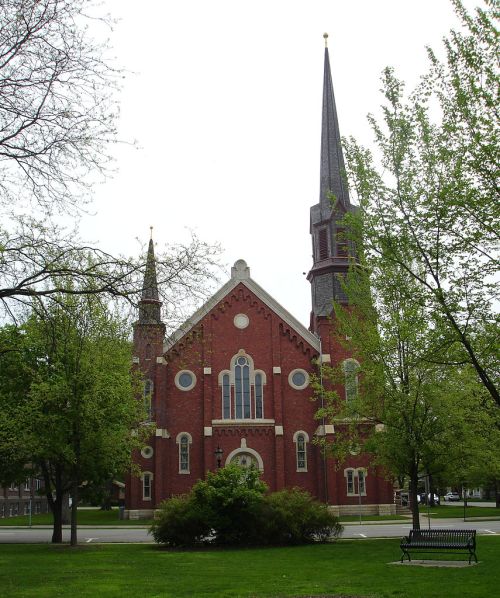 A red brick church with tall spires, surrounded by green trees and grass under a cloudy sky.