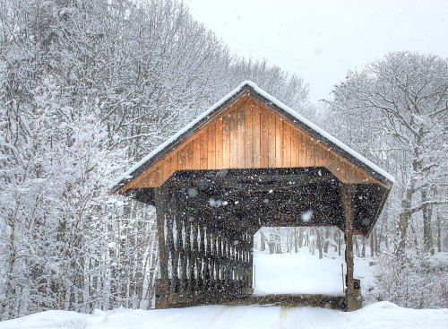 A snow-covered wooden covered bridge surrounded by trees in a winter landscape, with snowflakes falling gently.