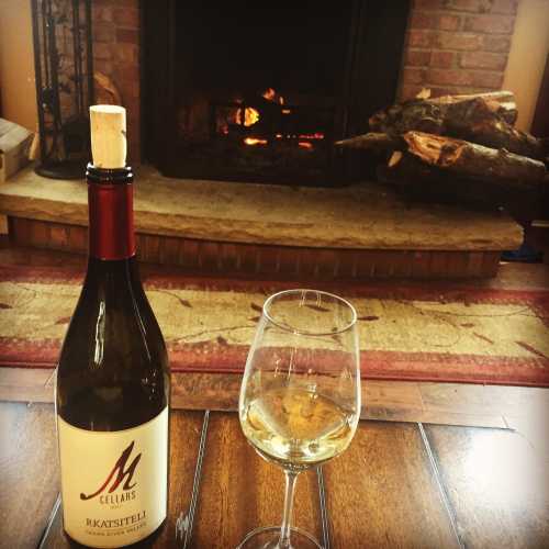 A bottle of white wine and a glass sit on a wooden table in front of a cozy fireplace with logs.