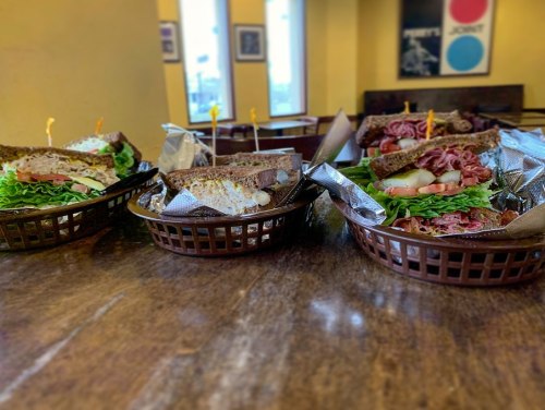 Four sandwiches in baskets on a wooden table, featuring various fillings and garnishes, with a cozy café atmosphere.