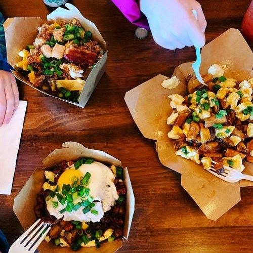 Three takeout boxes on a wooden table, filled with various dishes topped with green onions. Hands are reaching for the food.