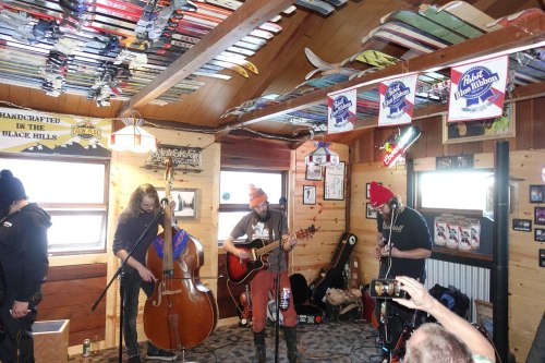 A band performs indoors with instruments, surrounded by vintage skis and beer signs in a cozy, rustic setting.