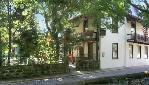 A charming two-story building surrounded by trees, featuring balconies and a stone wall along a quiet street.