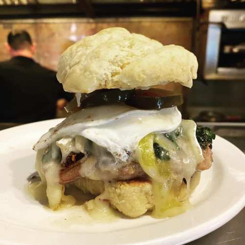 A hearty sandwich with a fluffy bun, layers of meat, cheese, spinach, pickles, and a fried egg, served on a plate.