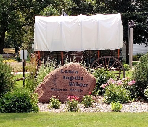 A covered wagon near a stone monument for the Laura Ingalls Wilder Memorial Society, surrounded by flowers and greenery.