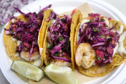 Three shrimp tacos topped with purple cabbage, pico de gallo, and lime wedges on a white plate.