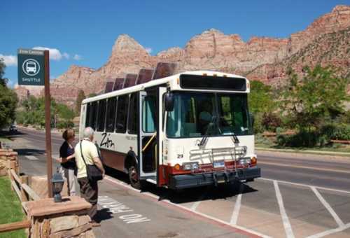 A shuttle bus parked near a sign for Zion Canyon, with scenic mountains in the background and two people waiting nearby.