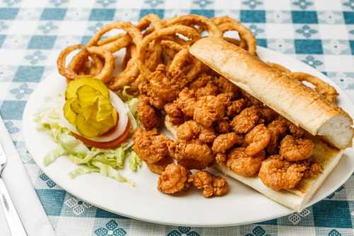 A plate of fried shrimp with a sandwich, onion rings, lettuce, tomato, and pickles on a checkered tablecloth.