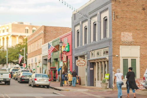 A bustling street scene with shops, people walking, and American flags in a historic downtown area.