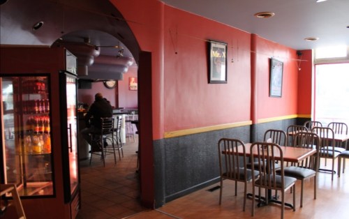 Interior of a casual restaurant with red walls, empty tables, and a bar area in the background.