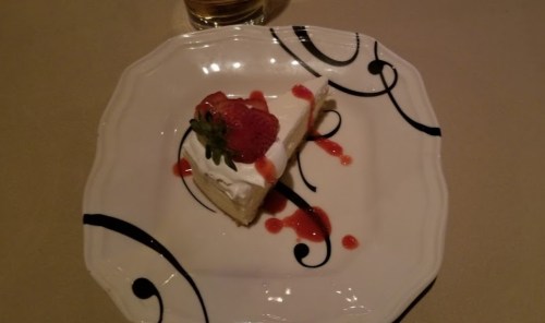 A slice of cheesecake topped with whipped cream and a strawberry, drizzled with strawberry sauce on a decorative plate.