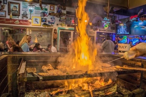 A vibrant fire blazes in a grill, surrounded by a lively restaurant atmosphere with patrons in the background.