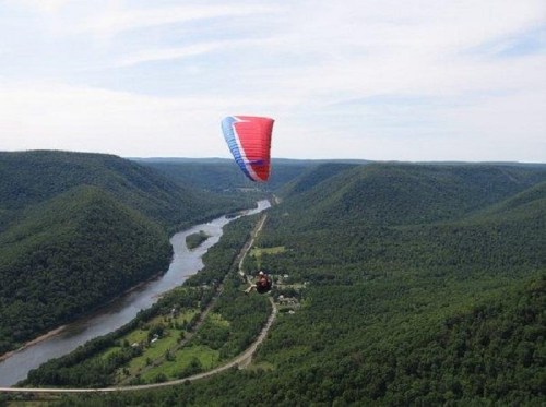 A paraglider soaring above lush green hills and a winding river under a clear blue sky.