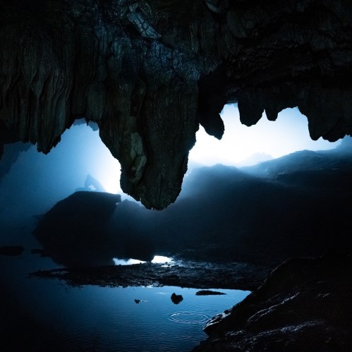 A dark cave interior with stalactites, illuminated by soft light, revealing a tranquil water surface.
