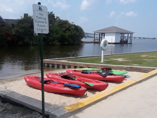 Red kayaks and canoes on a sandy launch area by a calm waterway, with a "No Parking" sign nearby.