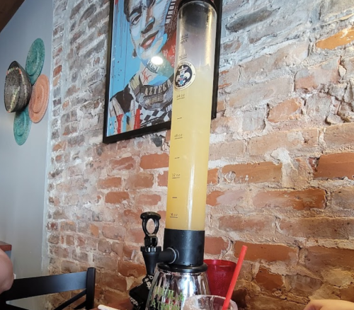 A tall drink dispenser filled with a yellow beverage, set against a brick wall and colorful artwork.