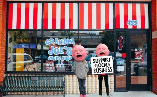 Two cartoonish figures with oversized heads stand outside a candy shop, holding a sign that says "Support Local Business."