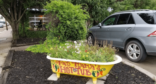 A colorful bathtub planter labeled "Fat Mama's Hot Tub" with flowers, parked car, and greenery in the background.