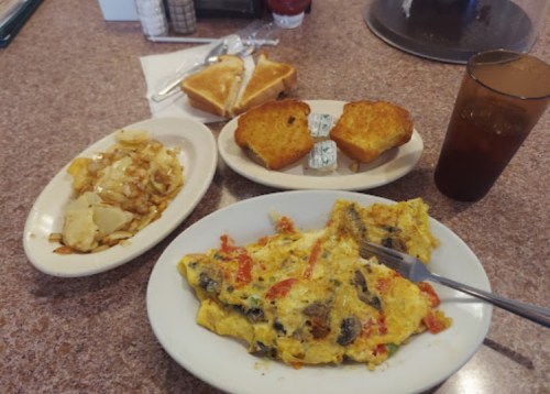 A plate of omelette with vegetables, hash browns, cornbread, and a drink on a diner table.